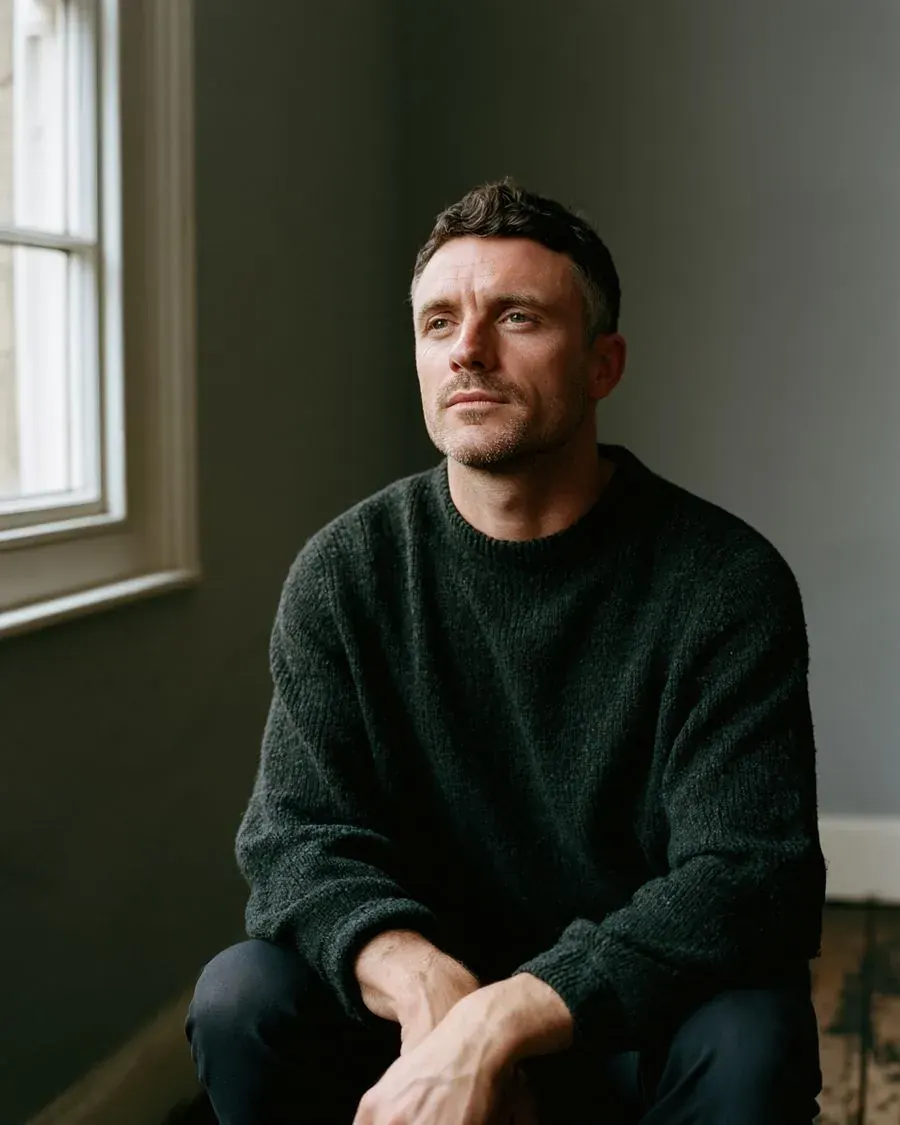 Daniel Mercer sitting beside a window in a plain room, in a dark green knit jumper, looking past the camera.