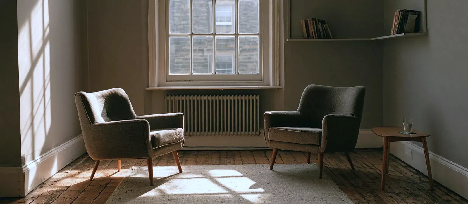 The consulting room: two armchairs facing each other across a rug, a sash window and a radiator between them, late afternoon light on the floorboards.